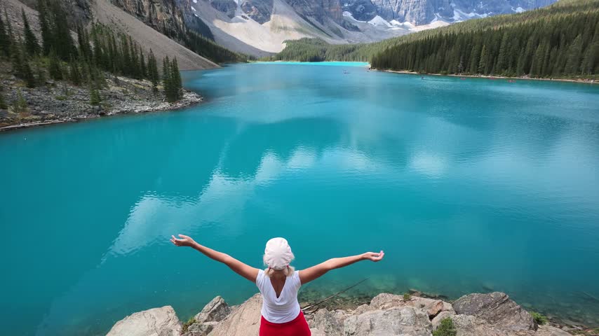aerial view: woman on rocks with arms outstretched, experiencing freedom and connection with nature, looking at stunning turquoise water, Lake Moraine reflecting in Banff National Park, Alberta,Canada
