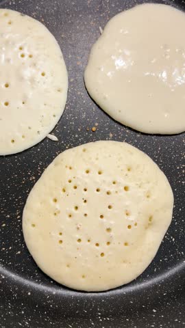Three pancakes cooking in a nonstick frying pan. Small bubbles appearing on the raw batter surface. Top view of homemade breakfast preparation in the kitchen. Sweet delicious. Vertical video.