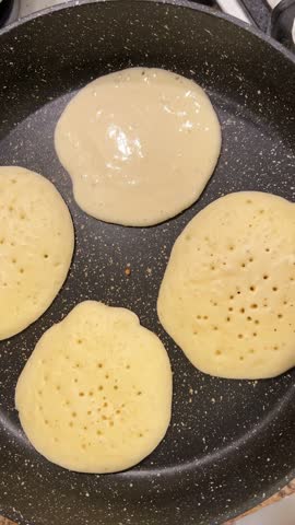 Cooking homemade pancakes on nonstick pan surface. Tiny bubbles form in raw batter while frying. The piece is flipped showing perfect golden brown crust. Metal spatula moves under dough for turning over. Vertical video.
