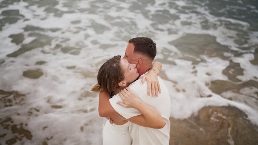 High-angle view of a loving couple in white clothing passionately hugging and kissing on a sandy beach, with gentle ocean waves washing over their feet during a romantic holiday getaway
