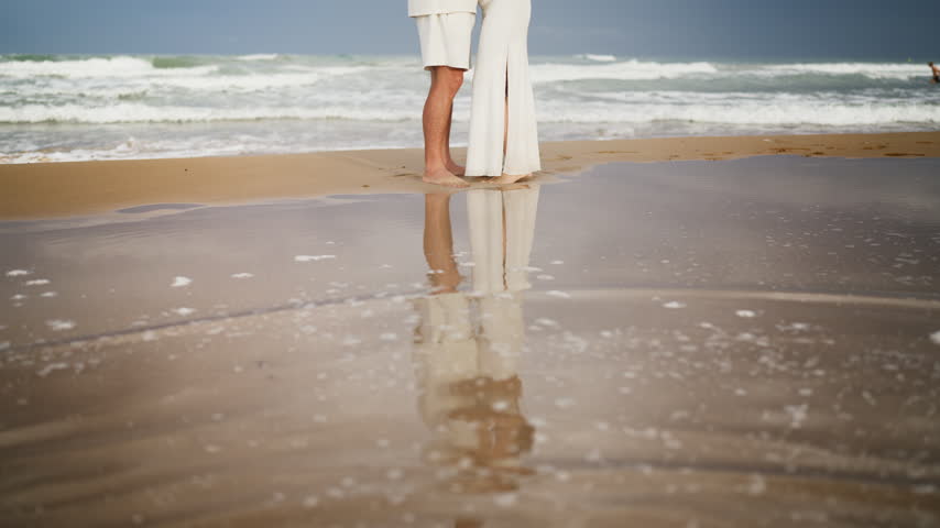 Romantic couple dressed in white standing barefoot together on the wet sand of a beach. Their reflection is visible in the shallow water as gentle ocean waves wash ashore in the background