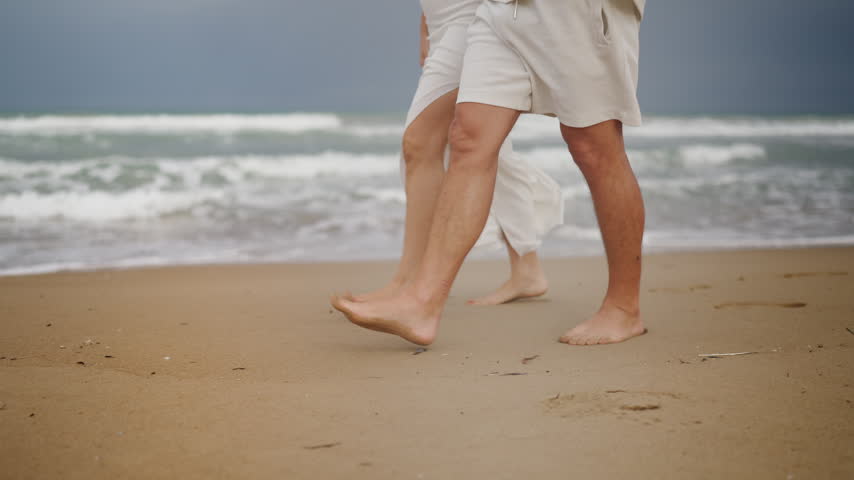 Close-up on the legs and feet of a man and woman wearing white clothes, walking barefoot together on a sandy beach while gentle ocean waves roll ashore during their vacation