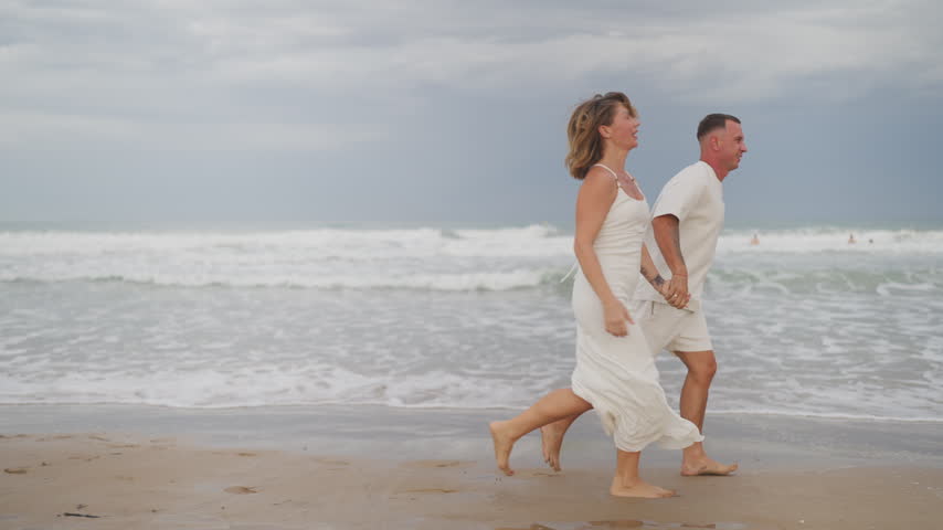Cheerful barefoot couple in white summer clothes laughing and running along the wet sand by the sea, holding hands and enjoying a romantic holiday together on an overcast day