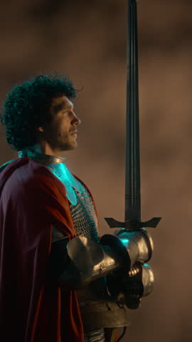 Vertical shot of young adult Caucasian man wearing knight armor holding sword upright. Clip fits fantasy gaming promotions, historical drama trailers, and cinematic advertising