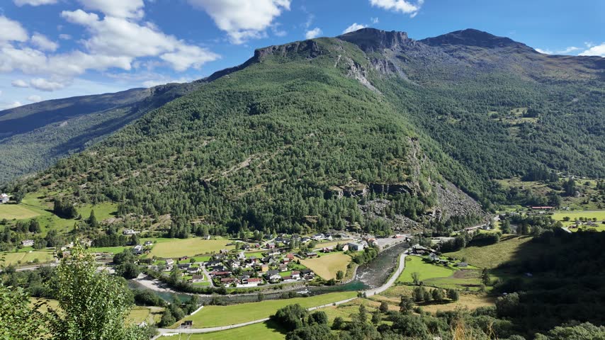 The view looking across the valley from the Brekkefossen viewpoint, just outside the small village of Flåm in rural Norway. Local homes are visible below, with the Flåmselvi river flowing through the centre of the shot. There is a dramatic tree-covered mountain in the background.