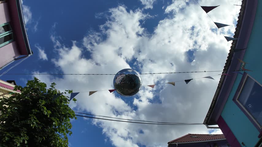 Walking underneath a large disco ball hanging outdoors between two buildings. There is multi-coloured bunting flags hanging across the street, and the buildings on either side are different colours.