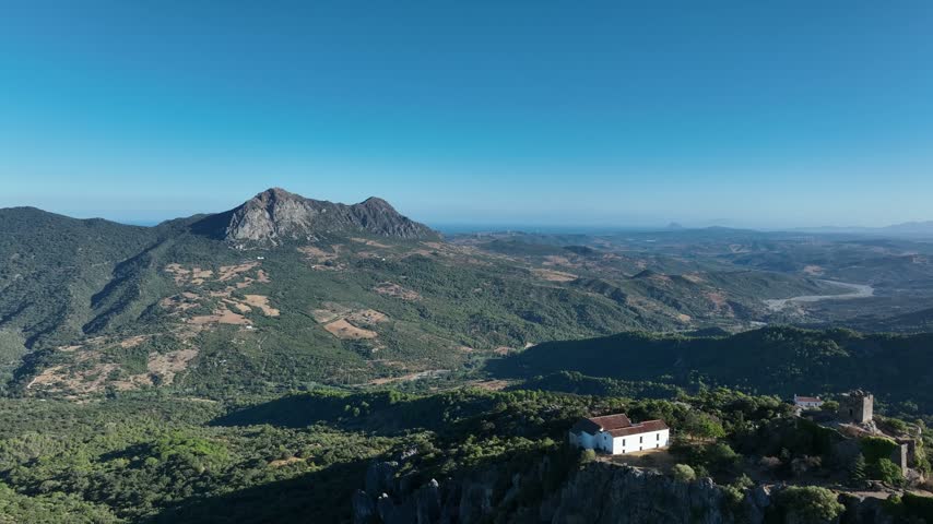 A stunning view of a mountain landscape shows a small white house placed amidst greenery. The scene captures the beautiful hills and blue sky.