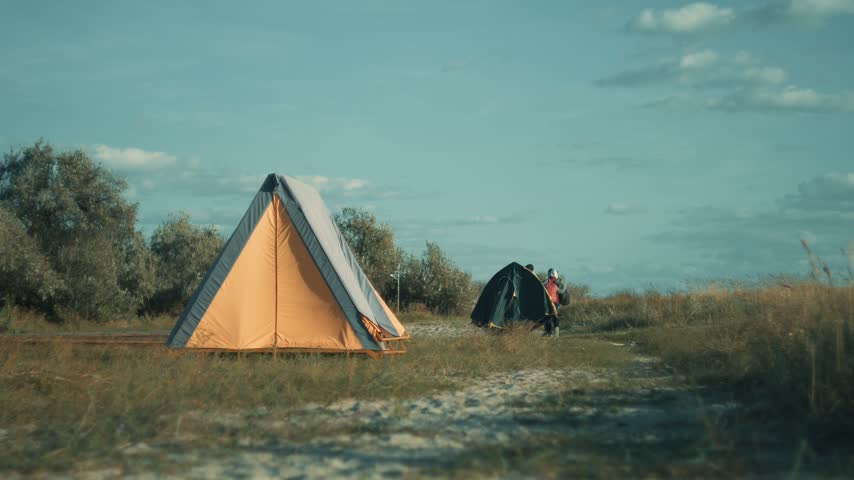 A vintage canvas tent stands on grassy beach dunes near trees, while a small group of campers carrying backpacks and gear walks toward a second tent further away. Summer weekend with friends on hikes.