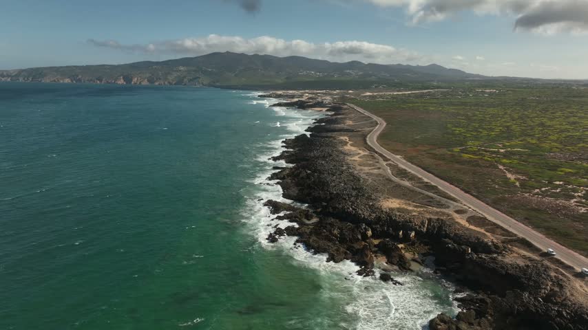 Footage captures a wide view of the coastline featuring rocky edges and crashing waves. A highway runs parallel to the shore, with hills visible in the distance under a clear sky.