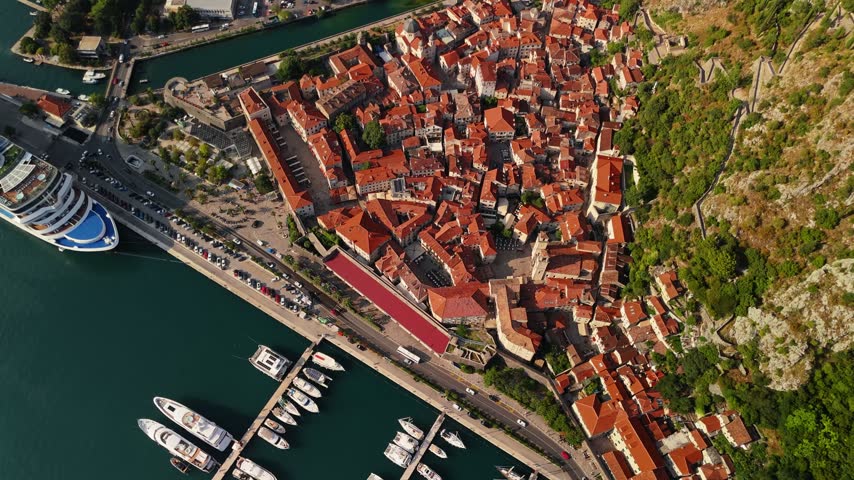 Kotor old town, Montenegro, in a top down aerial view. Medieval walls wrap the town while marina pontoons and a cruise ship line the waterfront below. Hard midday light throws sharp shadows across the roof ridges.