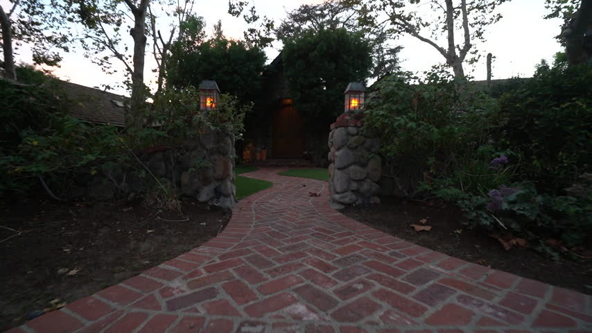 The camera moves along a curving herringbone brick walkway past lantern-topped stone pillars toward a stone house at twilight.