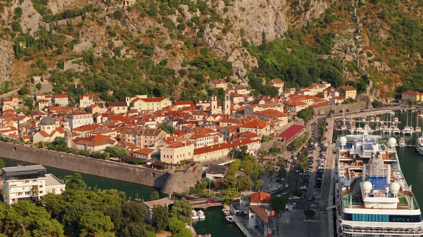 Kotor old town, Montenegro, ringed by stone defensive walls and a water moat. Marina pontoons, roads and a cruise ship fill the frame beyond the fortification. Warm afternoon light across a UNESCO listed town.