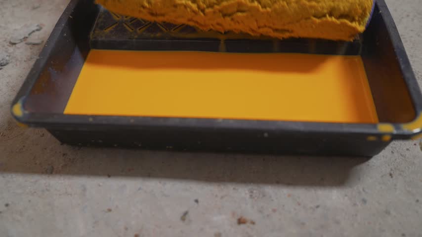 Close up video shows a paint roller being dipped and loaded with bright yellow paint in a black plastic tray. Preparation for painting, home renovation, or DIY project. Concrete floor background