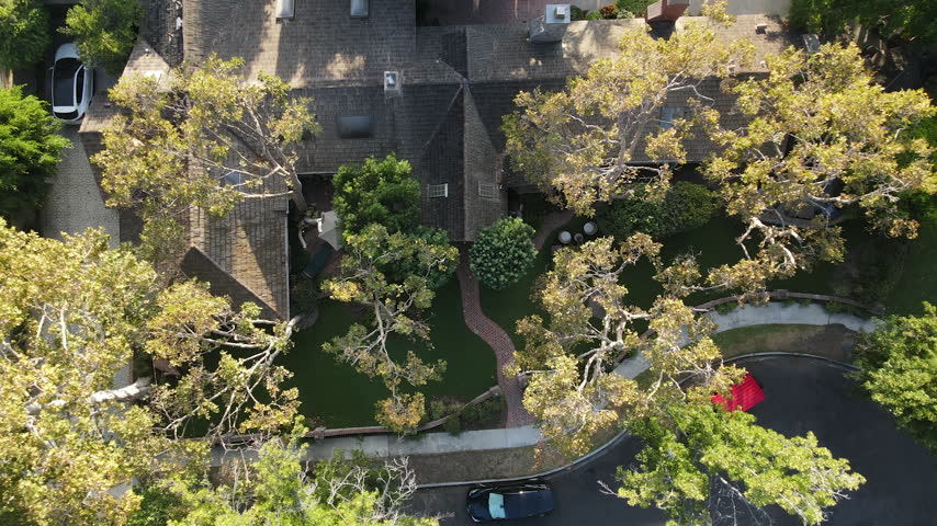 Top down drone view rises over a shingle roofed house, garden, backyard pool and a curving cul-de-sac with parked cars.