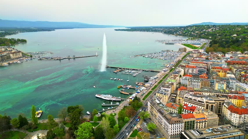 High angle cinematic drone footage capturing iconic Jet dEau fountain on Lake Geneva with vibrant urban scenery of Geneva city shoreline and surrounding mountains in background.