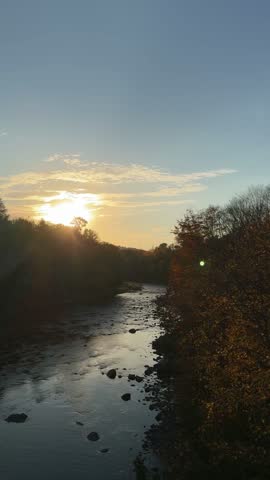 Vertical view of a river at golden sunset, surrounded by autumn forest and colorful fall foliage, with warm sunlight reflecting across the water and rocks visible in the stream.