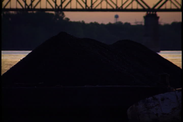 Zoom out from coal barge to Ohio River railroad bridge and power plant in silhouette at sunset in Metropolis, Illinois.