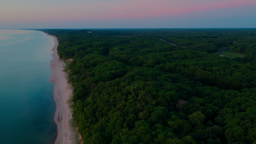 Lake Michigan shoreline at twilight, where dense hardwood forest meets a pale sand beach and calm shallow water under a soft pink sky.