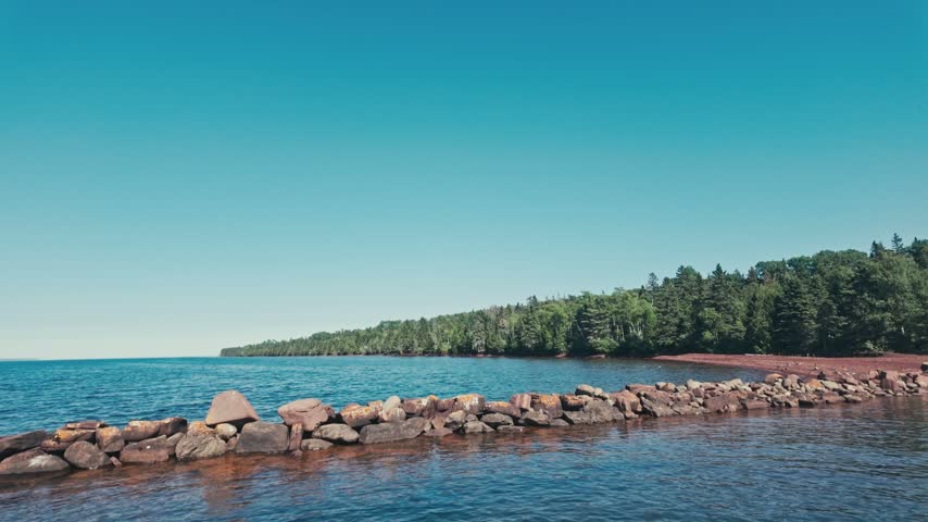 A panning shot of Lake Superior, a tall green tree line, and a rock break wall on a sunny day. Filmed at Isle Royale National Park.