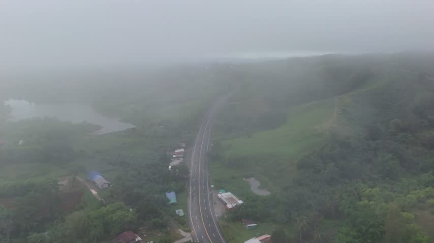 Aerial drone footage captures winding road through vibrant mountain landscape with serene lake under heavy fog and low clouds.