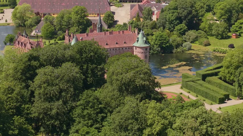 A telephoto aerial view gradually reveals Egeskov Castle through dense green garden foliage, creating a layered composition of historic architecture and nature.