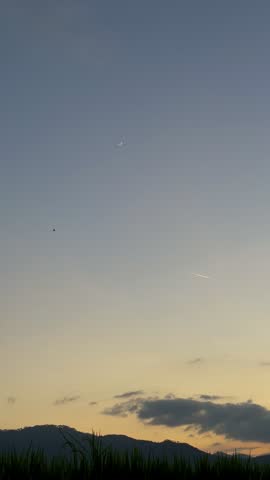 Vertical video of a bright crescent moon and star aligned in the evening twilight sky over dark hills silhouette in Bali. An airplane contrail moves softly across the glowing sky above clouds at dusk.
