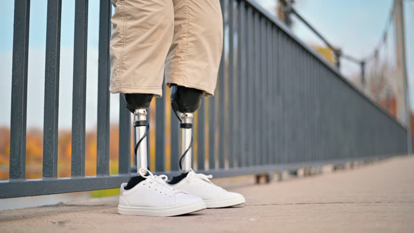 Close-up view of person with prosthetic legs, moving slowly, standing on bridge railing. Autumn scenery in background.