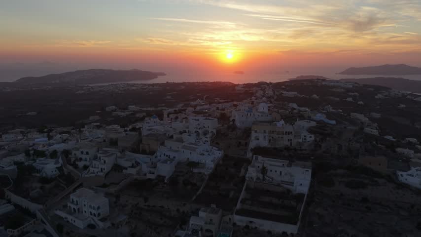 Cinematic drone shot flying over Santorini village during golden hour with sun setting over Aegean sea horizon.