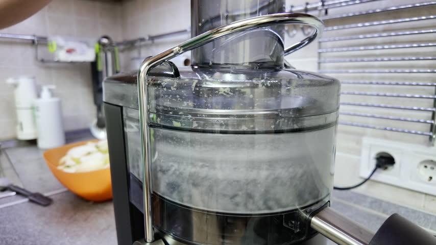 Close-up static shot of a modern electric juicer processing peeled green apple slices inside a transparent chute. A human hand appears on the background blurred edge near an orange bowl under neutral indoor lighting.