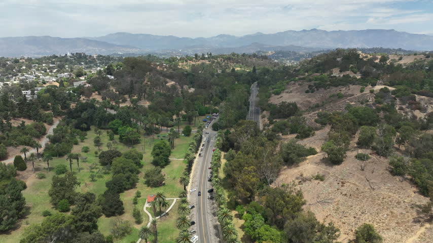 Los Angeles, California, USA - Aerial drone view over an Elysian Park picnic area with green hillsides, trees, and a mountain range visible in the distance. The footage captures the urban park