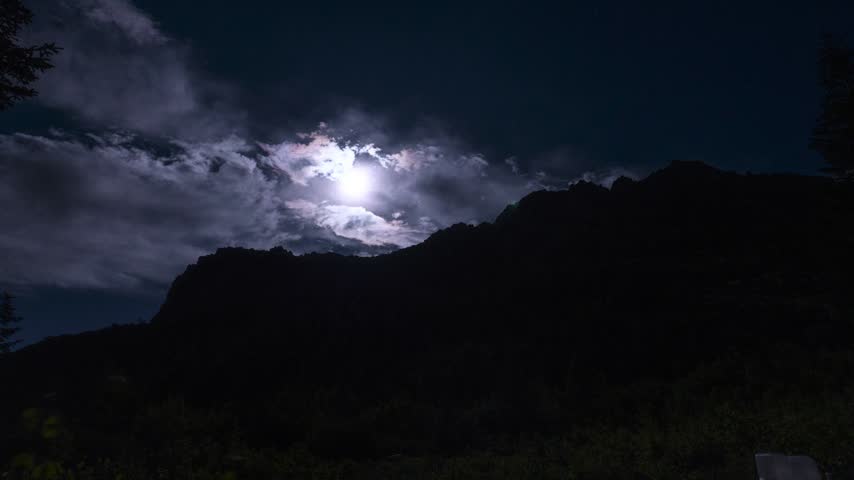 Bright moon emerges through drifting clouds above a rugged mountain ridge and dark forest at night.