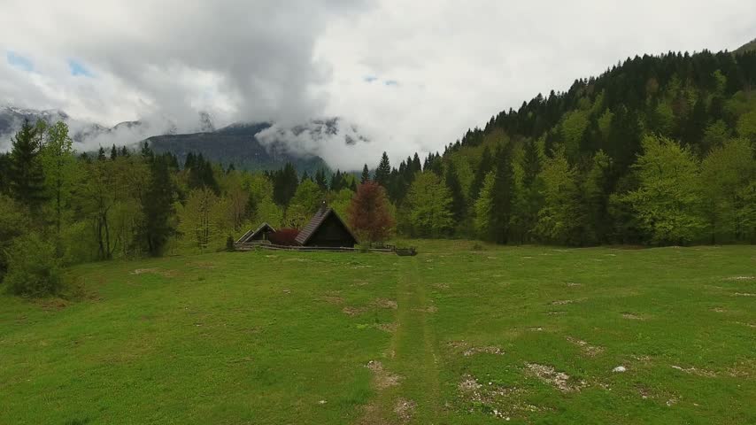 Aerial view of a small wooden cabin nestled in a lush green meadow near Lake Bohinj, surrounded by dense forests and misty mountains in Bohinjsko jezero, Radovljica, Slovenia.