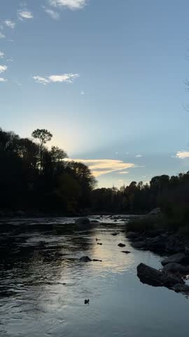 Vertical view of a flowing forest river at sunset, with dark tree silhouettes, rocks, calm ripples and warm evening light reflecting across the water under a blue sky.