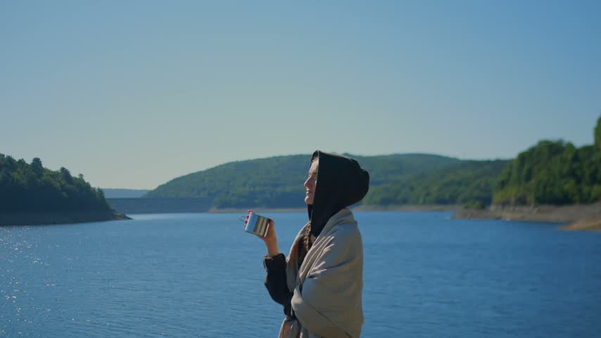 Woman holding a cup beside the calm waters of Colebrook River Lake, CT, surrounded by forested hills, conveying mindfulness, peace, relaxation, and connection with nature. 4K.