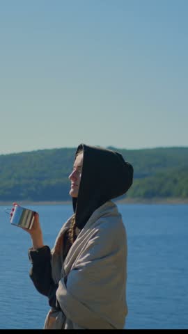 Woman holding a cup beside the calm waters of Colebrook River Lake, CT, surrounded by forested hills, conveying mindfulness, peace, relaxation, and connection with nature. 4K. Vertical.