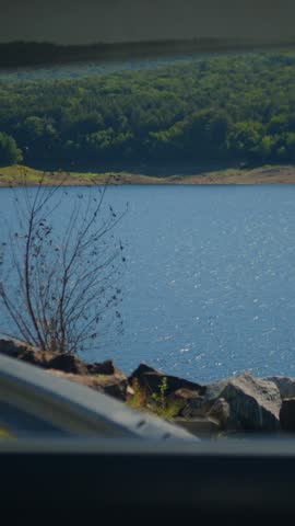 Scenic view of Colebrook River Lake in Connecticut, USA, seen through a car window with a rocky shore, roadside guardrail, and forested hills.