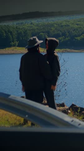 Couple embracing along the shore of a sunny reservoir, viewed through a car window, with blue water, rocks, a guardrail, and forested hills.