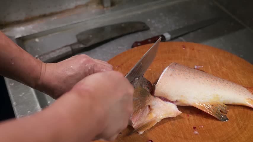 Close up shot of professional chef hands carefully slicing and filleting a fresh raw fish on a round wooden cutting board in a commercial kitchen setup.
