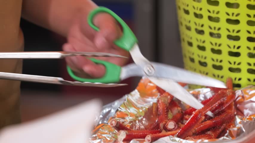 A close-up shot capturing a street food vendor or chef using kitchen scissors and metal tongs to cut cooked, bright red king crab legs into pieces over aluminum foil.