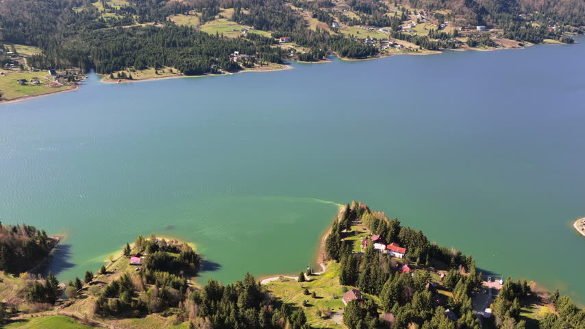 Aerial drone view of a forested lakeshore with houses partially hidden among trees, sunlight reflecting on the water, at Colibita Lake, Romania