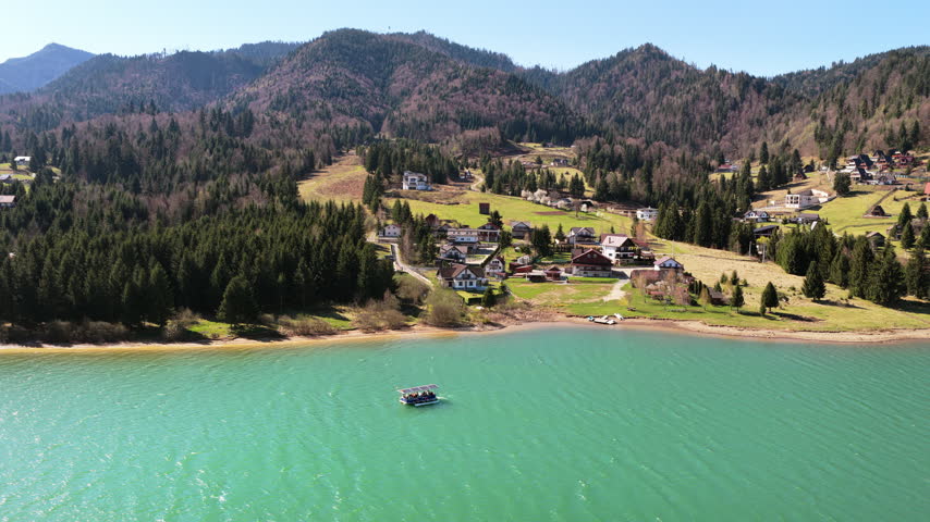 Aerial drone view of a small lakeside settlement along Colibita Lake, with wooden houses, green hills, and pine forests