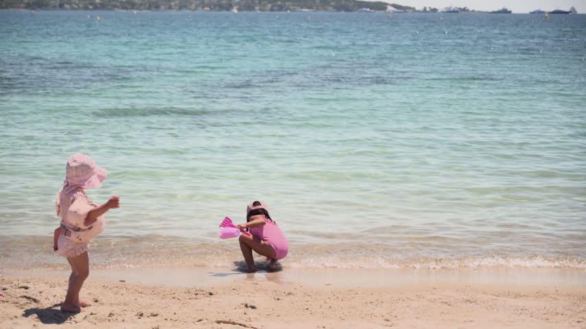 Two young children in pink swimwear playing at the shoreline of a calm sandy beach on a sunny summer day