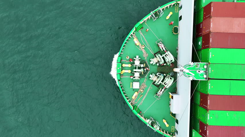 multiple ship crew members in bright safety orange working with massive ropes and heavy machinery on the vibrant green deck of a large vessel
