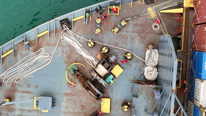 multiple ship crew members in bright safety orange working with massive ropes and heavy machinery on the vibrant green deck of a large vessel