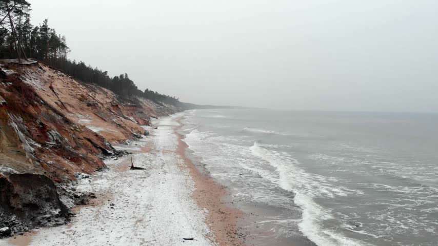 Lone person walks on sandy shore beneath steep red clay cliffs along icy coastline under moody overcast winter sky footage.