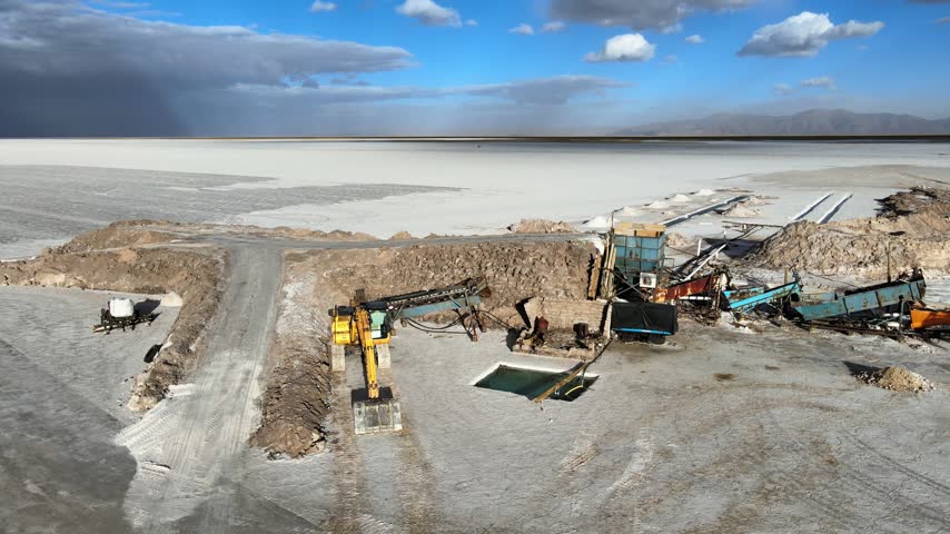 Industrial lithium mining machinery sits on the Salinas Grandes salt flat under bright daytime skies, with excavators, conveyors and pools framed by the vast white desert landscape.