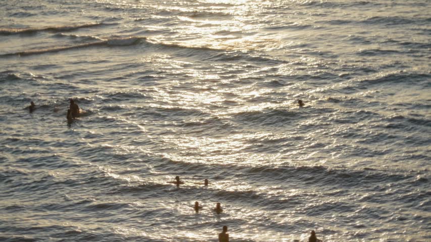 Silhouettes of people bathing and enjoying the gentle waves of the Mediterranean Sea in Tel Aviv, Israel, as the setting sun creates a shimmering golden reflection on the calm water surface