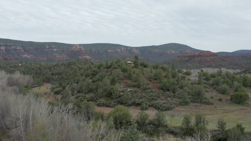 Aerial view captures rolling hills covered in evergreen trees and red sandstone rock formations under cloudy sky in Sedona Arizona.