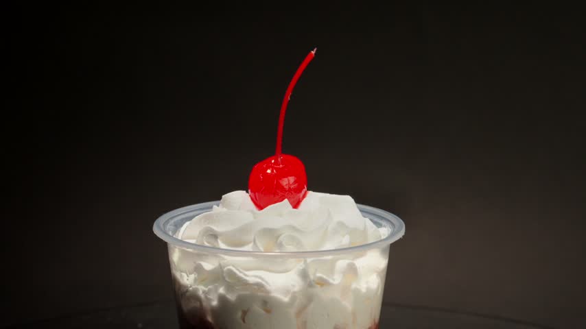 Close up rotation shot of delicious chocolate pudding dessert topped with whipped cream and maraschino cherry in plastic cup.