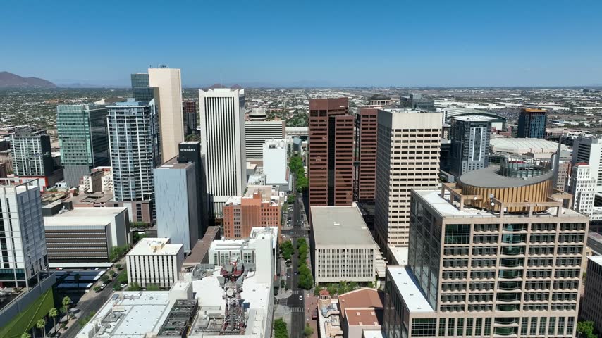 Aerial drone footage showcasing urban sprawl of downtown Phoenix Arizona with contemporary skyscrapers parking structures and city streets under bright sunny clear blue sky during daylight hours.